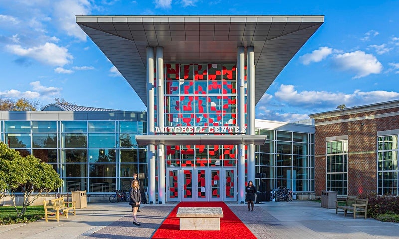 Front view of the Mitchell Recreation & Athletic Center main entrance with large glass windows, red accents, and a red carpet leading to the entrance under a modern overhang.