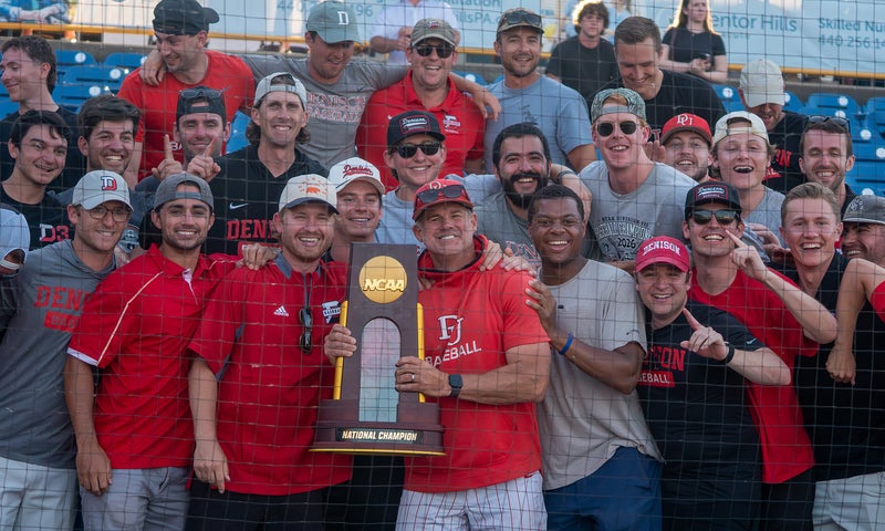 A group of Denison alumni, many wearing red shirts and caps, gather and smile around Coach Mike Deegan in the center holding a championship trophy. 