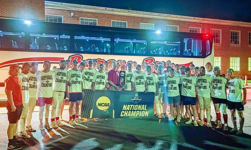A group of people in matching shirts stand outside at night in front of a bus and building, holding a large banner that reads NCAA National Champion.