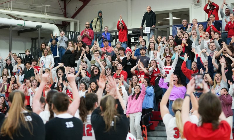 A crowd of fans fill the bleachers as women's basketball players salute them from the court.