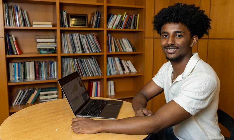 A person sits at a round table in front of a laptop, smiling, with bookshelves filled with books in the background.
