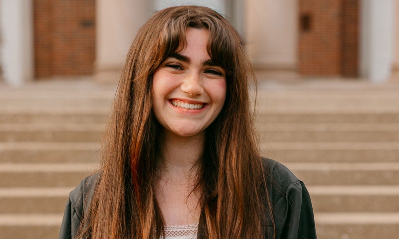 A young woman with long brown hair smiles at the camera while standing outdoors in front of stone steps.