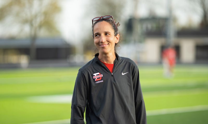 A woman with dark hair in athletic gear stands outside on a sports field.