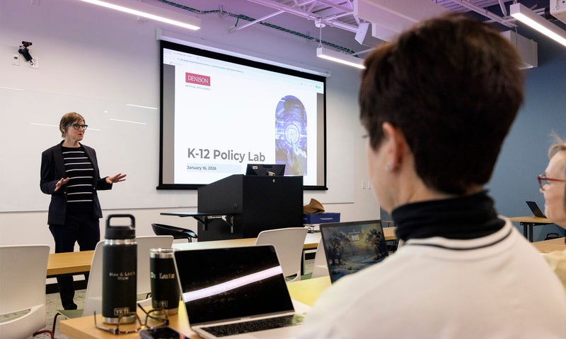 A person stands and presents in front of a projected slide titled "K-12 Policy Lab" to an audience seated with laptops in a classroom.