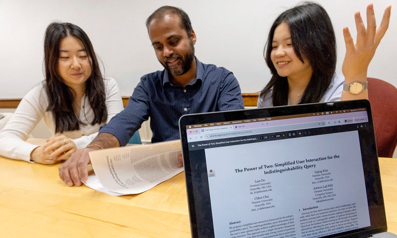 Two college students and a faculty member sit at a table discussing a printed document, which is digitally presented on a laptop screen in the foreground.
