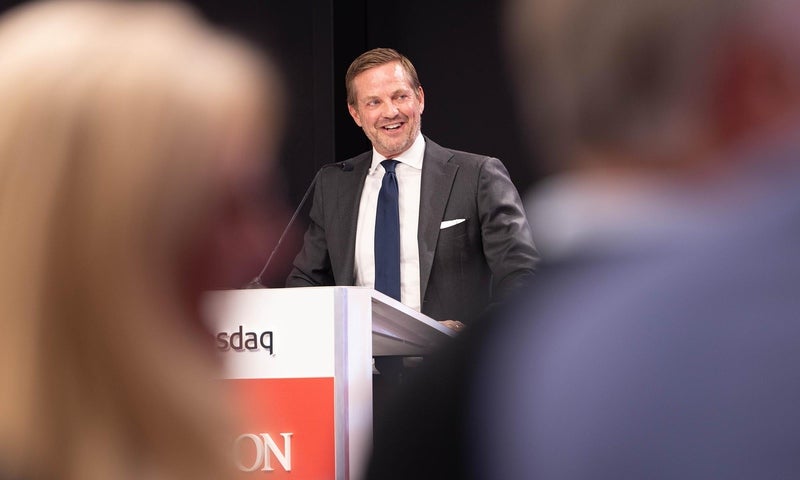 A man in a suit and tie stands at a podium, speaking into a microphone, with a smile on his face. The foreground is blurred, showing parts of two people facing him.