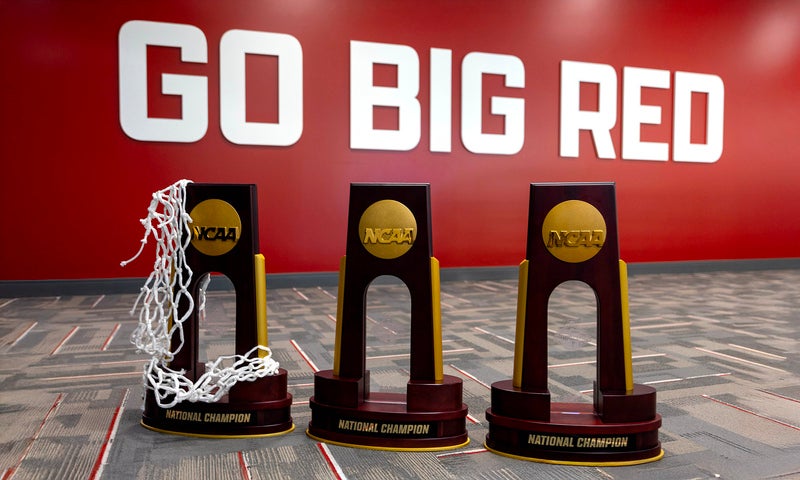 Three NCAA championship trophies lined up in front of a large red well with the white block letters that say "Go Big Red"