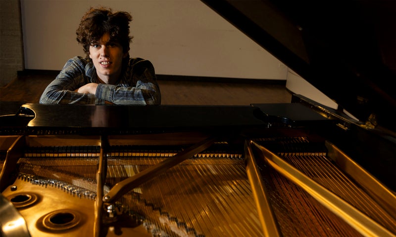 A person with curly hair leans on a grand piano, looking toward the camera. The pianos strings and internal components are visible in the foreground.