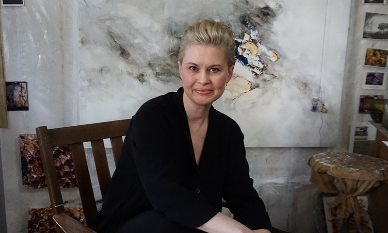 Allison Leigh, a woman with blonde hair, sits on a wooden chair in front of an abstract painting, wearing a black outfit. Artworks and materials are visible in the background.