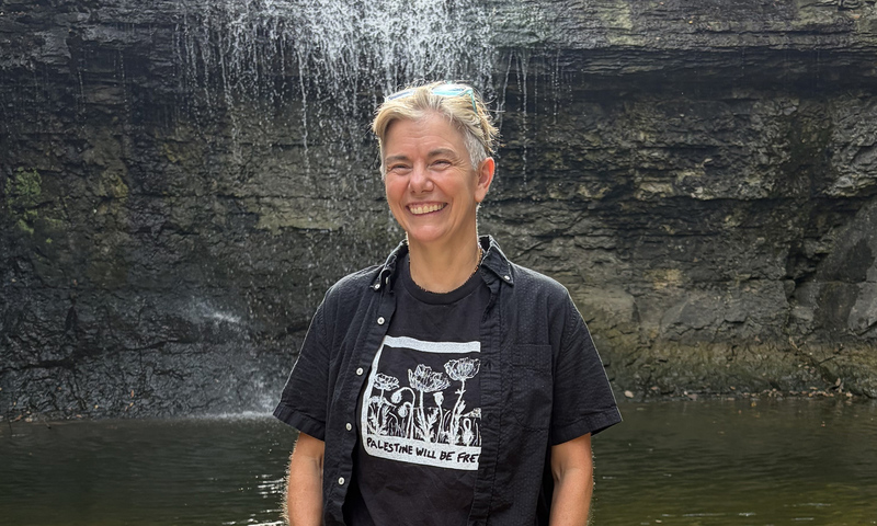 Person standing and smiling in front of a small waterfall, wearing a black shirt and sunglasses on their head.