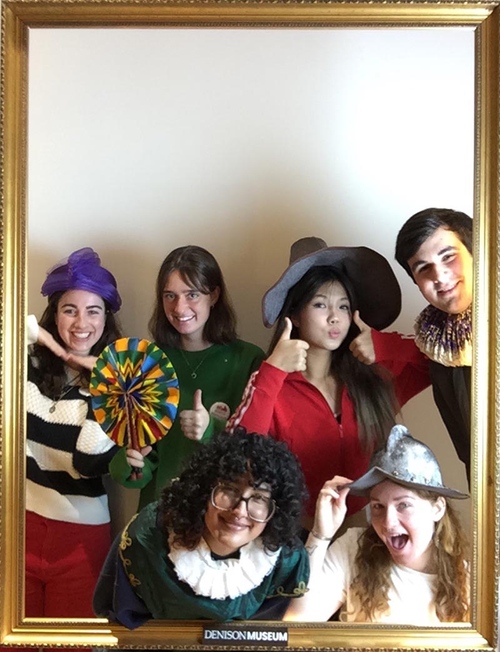 A group of Museum interns pose for a picture in a giant photo booth; the photo is bordered by a gold picture frame which displays a small sign along the bottom edge that reads "Denison Museum."
