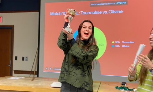 A student standing in front of a presentation projected on a screen holds up a trophy in celebration.