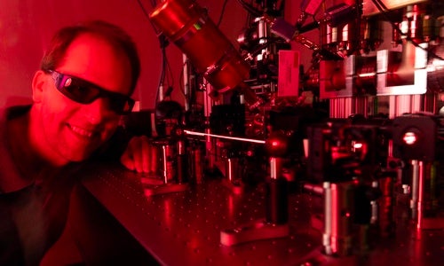 A man in dark protective glasses smiles next to a scientific piece of equipment emitting a laser in a red-hued room.