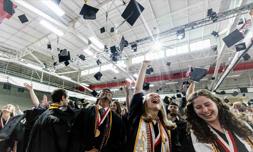 A large group of college graduates in caps and gowns toss their caps in the air in celebration in a large fieldhouse during a graduation ceremony.