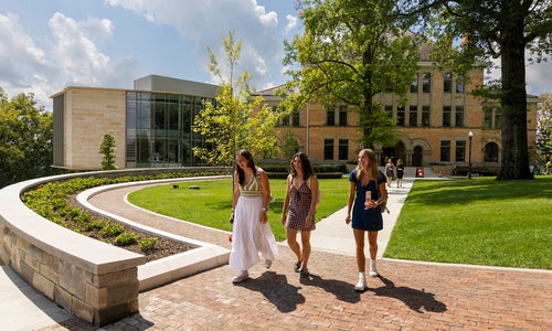 Three people walk along a brick path in front of a modern academic building on a sunny day, surrounded by green lawns and trees.
