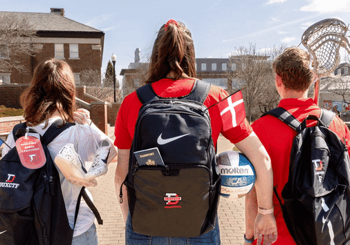 -Three athletes walking across the quad holding athletic gear. Their backpacks have flags sticking out of it that move slightly with the wind.