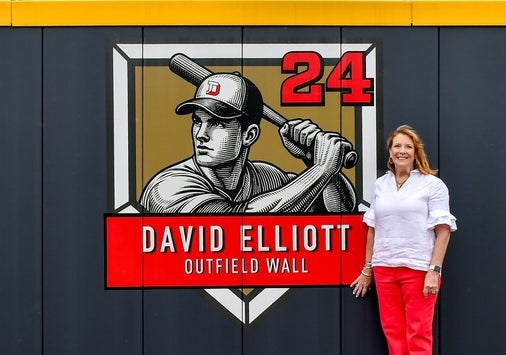 A photo of Lisa Elliott standing by the newly unveiled David Elliott outfield wall graphic.