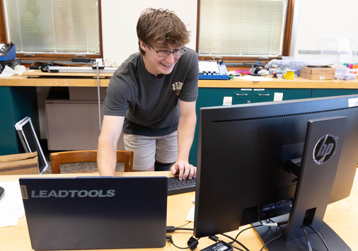 Photo of Denison student Joe Teates ’28 standing at a lab table, looking at computer monitors.