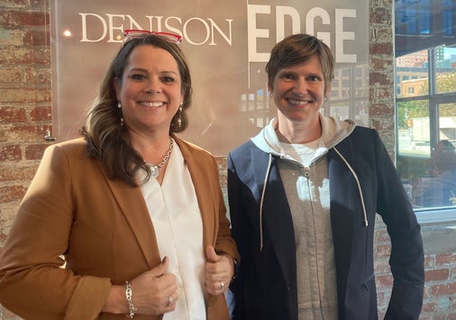 Two people, Kristi Wellington-Baker, Amazon Web Services Higher Education Strategy and Liv Gjestvang, Denison vice president and chief information officer, smiling and posing indoors in front of a sign that reads “DENISON EDGE,” with brick walls and windows in the background.