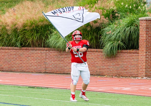A football player in a red and white uniform stands on a football field waving a large flag bearing the Mission 34 logo.