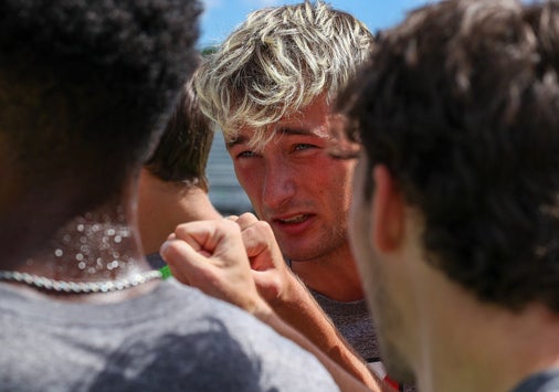 A close up photo of a group of college soccer players on the field; the face of a young man with blond hair is in focus in the center of the photo, surrounded by his teammates giving a celebratory fist bump.