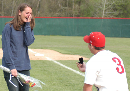 A baseball player in uniform kneeling and offering a wedding ring to a woman standing on a baseball field holding flowers.