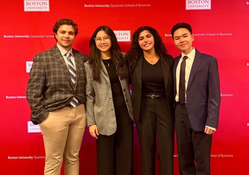 Four Denison students in business attire stand in front of a large red backdrop dotted with Boston University and BU Questrom School of Business logos ahead of their competition.