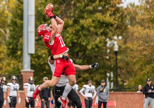 A football player in a red uniform jumps to catch a pass while a defender in a white and black uniform attempts to block him. Other players and trees are visible in the background.