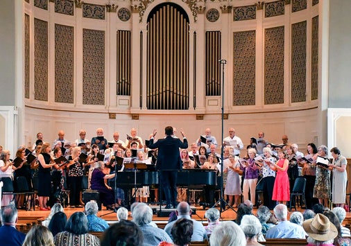 A choir and musicians perform on stage in a church or concert hall, directed by a conductor. The audience is seated in rows facing the performers. The background features a large organ and decorative architectural details.