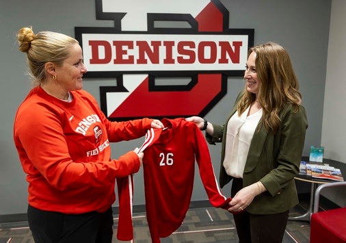 Two women stand indoors in front of a large Denison University logo. One hands a red sports jersey with the number 26 on it to the other. Both are smiling.