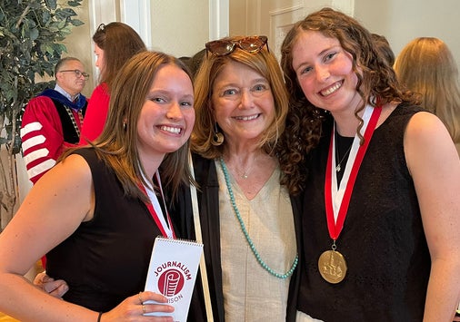 Three women stand indoors, smiling at the camera. Two younger women wear medals on red and white ribbons around their necks, and the other woman stands between them. Other people are visible in the background.