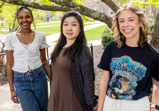 Three young women stand outdoors on a sunny day, posing and smiling in front of a leafy tree and campus buildings.