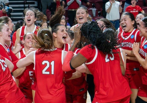 A womens basketball team in red uniforms celebrates together on the court, laughing and cheering. Player number 21 is visible in the center of the group.