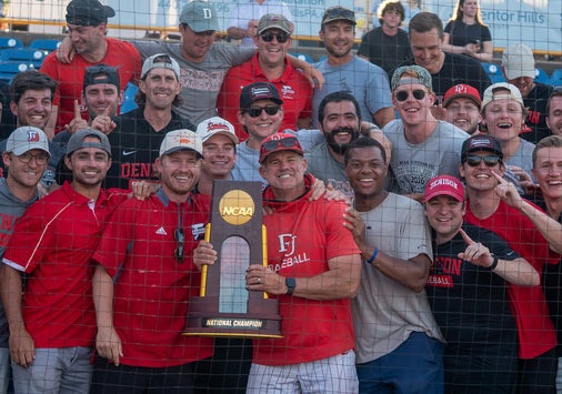 A group of Denison alumni, many wearing red shirts and caps, gather and smile around Coach Mike Deegan in the center holding a championship trophy. 