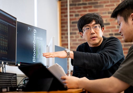 Two people work at a desk with computer monitors and a tablet, discussing data displayed on the screens in a modern office with exposed brick walls.