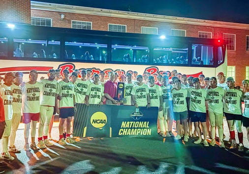 A group of people in matching shirts stand outside at night in front of a bus and building, holding a large banner that reads NCAA National Champion.