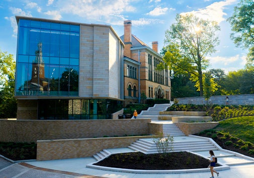 Modern glass and brick building with large windows, outdoor steps, and landscaped greenery under a partly cloudy sky.