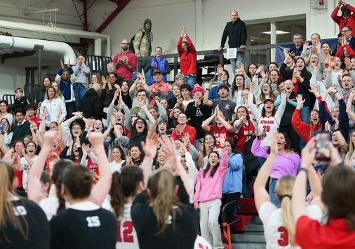 A crowd of fans fill the bleachers as women's basketball players salute them from the court.