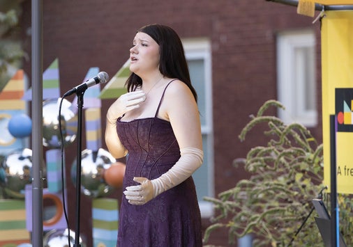 A woman with dark hair, wearing a purple sleeveless dress with straps, sings on an outdoor stage.