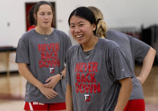 A women's basketball players in a Denison tee shirt that says "Never back down" smiles at practice with other players in the background.