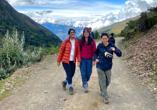 Three people on a hiking on a high-altitude trail with snow-capped mountains and clouds in the distance behind them.