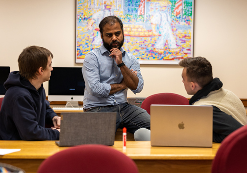 A man sits on a desk speaking with two students in a classroom with computers and colorful artwork on the wall in the background.