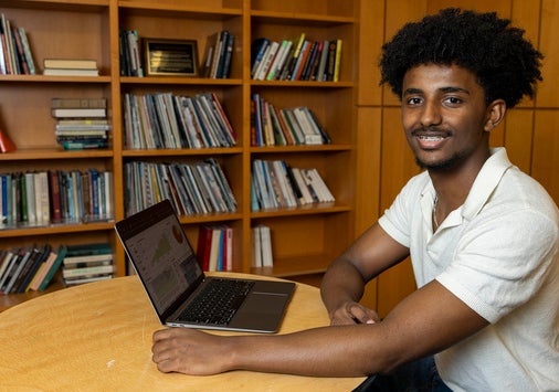 A person sits at a round table in front of a laptop, smiling, with bookshelves filled with books in the background.