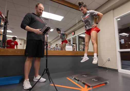A woman in athletic gear performs a vertical jump test while a man in sportswear records results on a tablet. The scene takes place in a gym with mirrors and exercise equipment visible in the background.