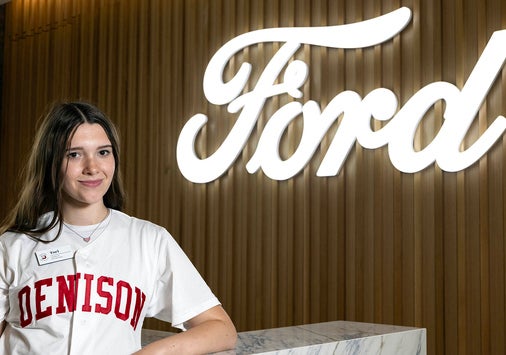 A person in a Denison t-shirt stands in front of a large illuminated Ford logo on a wood-paneled wall.