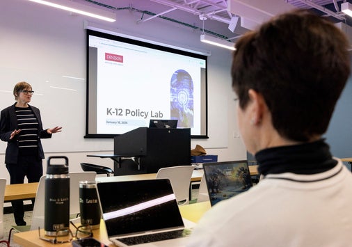 A person stands and presents in front of a projected slide titled "K-12 Policy Lab" to an audience seated with laptops in a classroom.