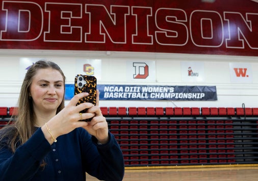 A person uses a phone to record a video inside a gymnasium with DENISON in large letters on the wall and a banner for the NCAA DIII Women's Basketball Championship in the background.
