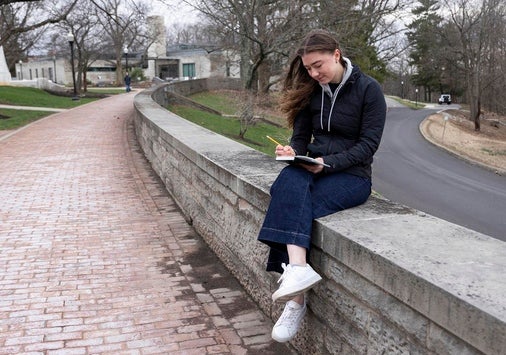A young woman sits on a curved stone wall along a brick path writing in her notebook.