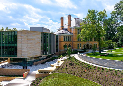 A modern building extension with glass and stone sits beside a large historic brick house, surrounded by green lawns, trees, and landscaped pathways under a partly cloudy sky.