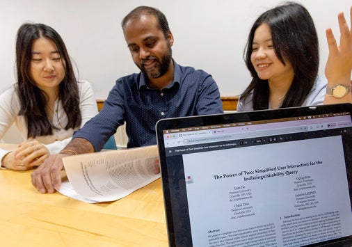 Two college students and a faculty member sit at a table discussing a printed document, which is digitally presented on a laptop screen in the foreground.